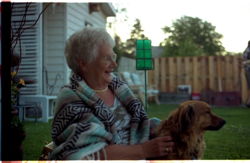 An older woman sitting outside her house with a small brown dog at her feet